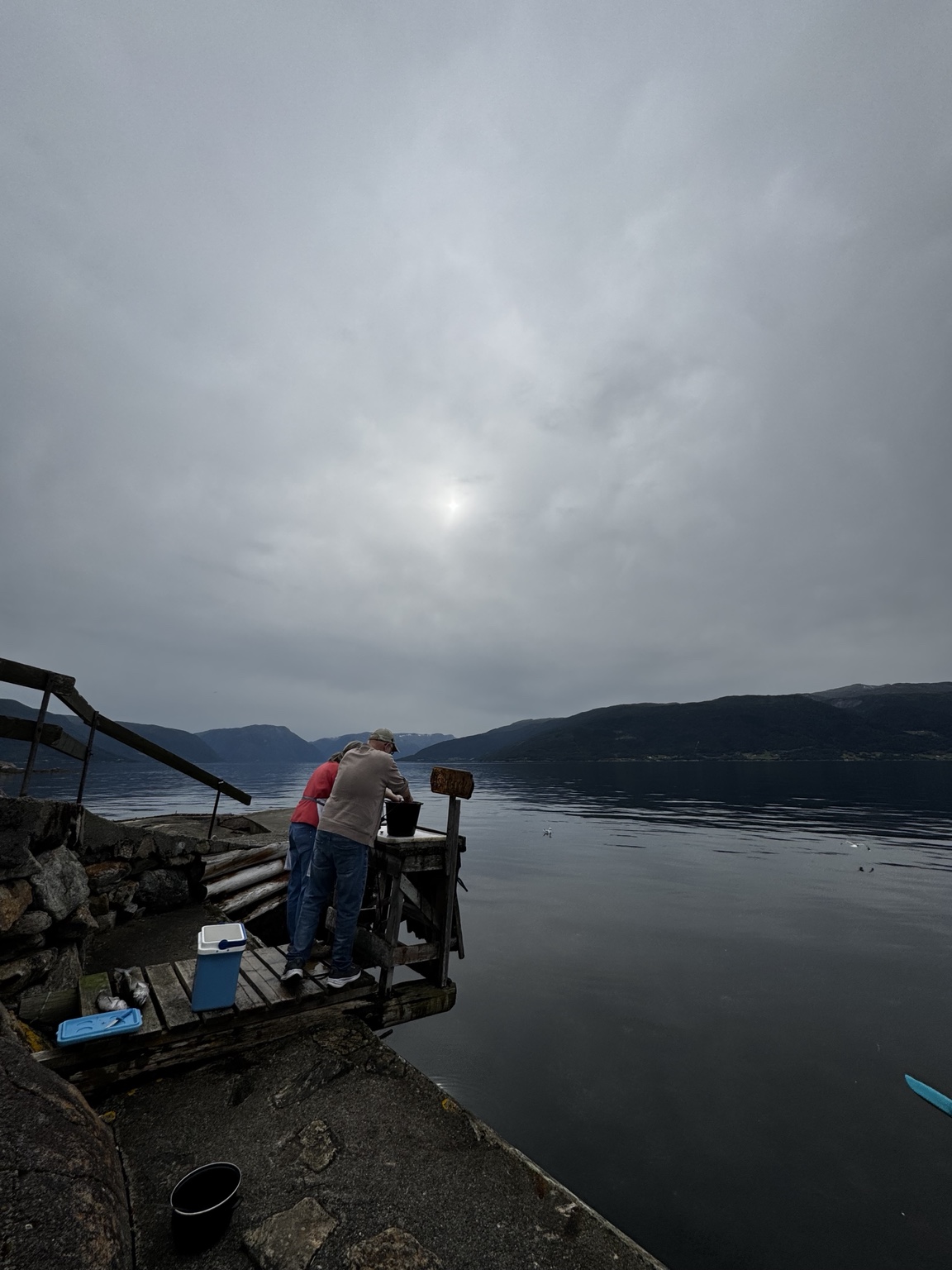 Foto van twee mensen op een houten steiger bij Tveit, met het fjord en een bewolkte lucht.