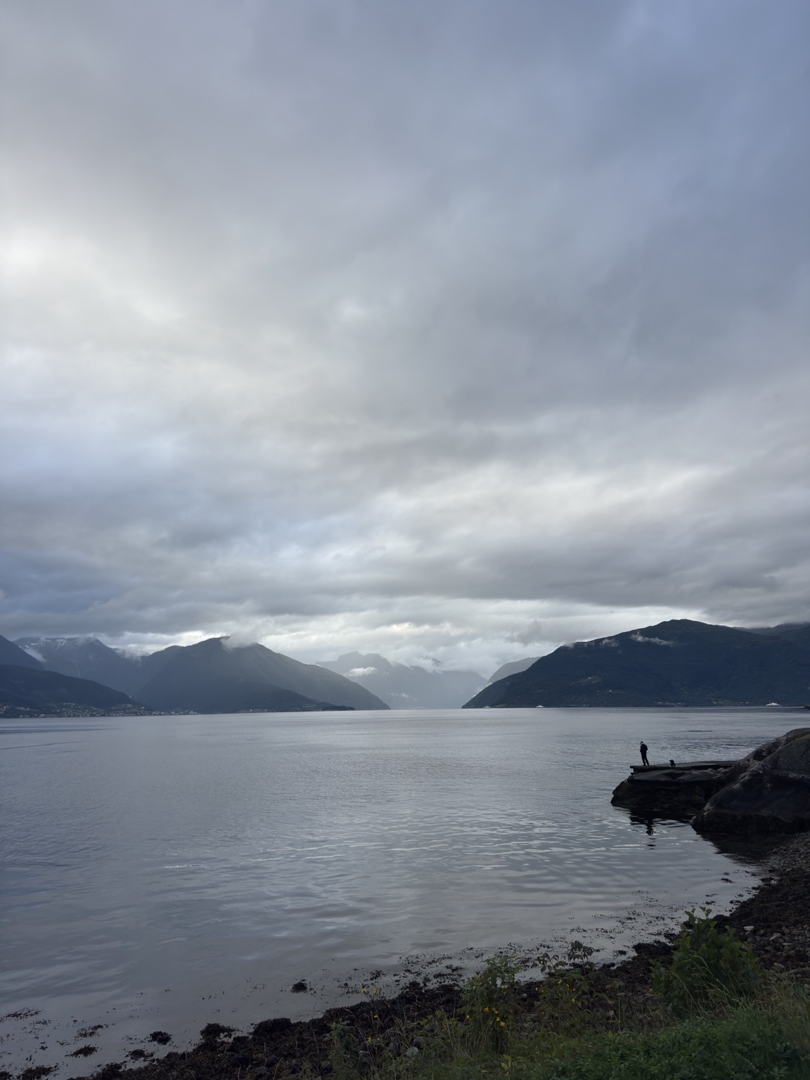 Foto van het Sognefjord vanaf Tveit, met bergen onder een bewolkte lucht en een persoon op de steiger.